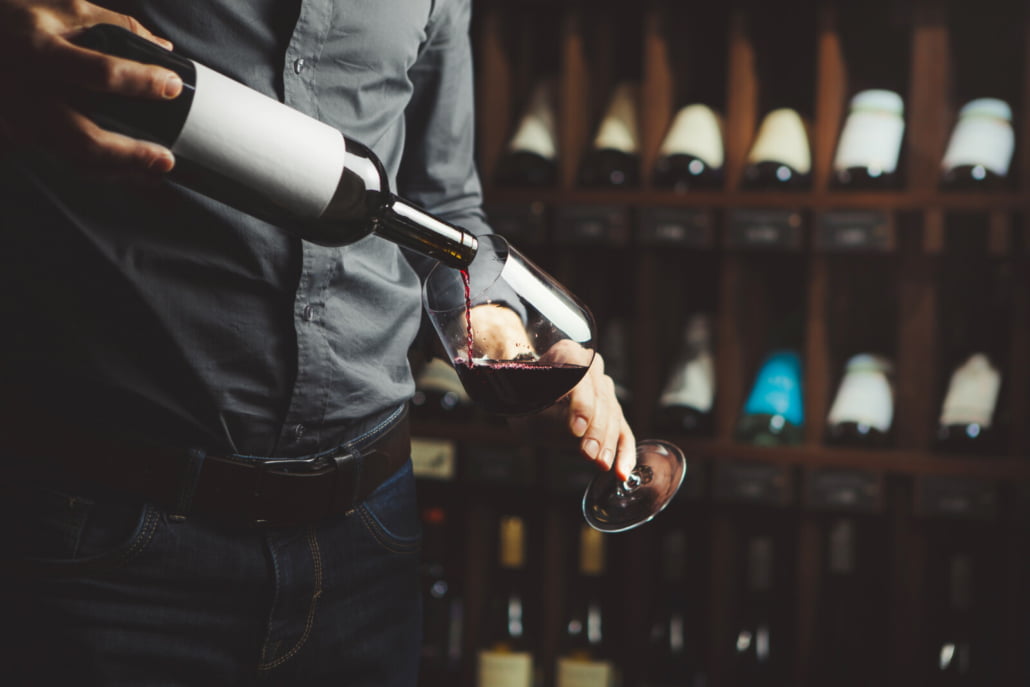 Close up shot of sommelier pouring red wine from bottle in glass on underground cellar background